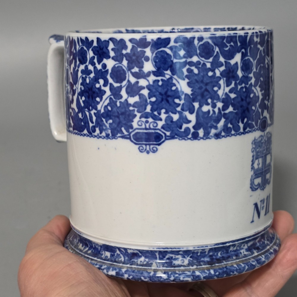 A cylindrical blue and white transferware ceramic tankard featuring a floral upper border and a coat of arms crest with the text No 11. View X