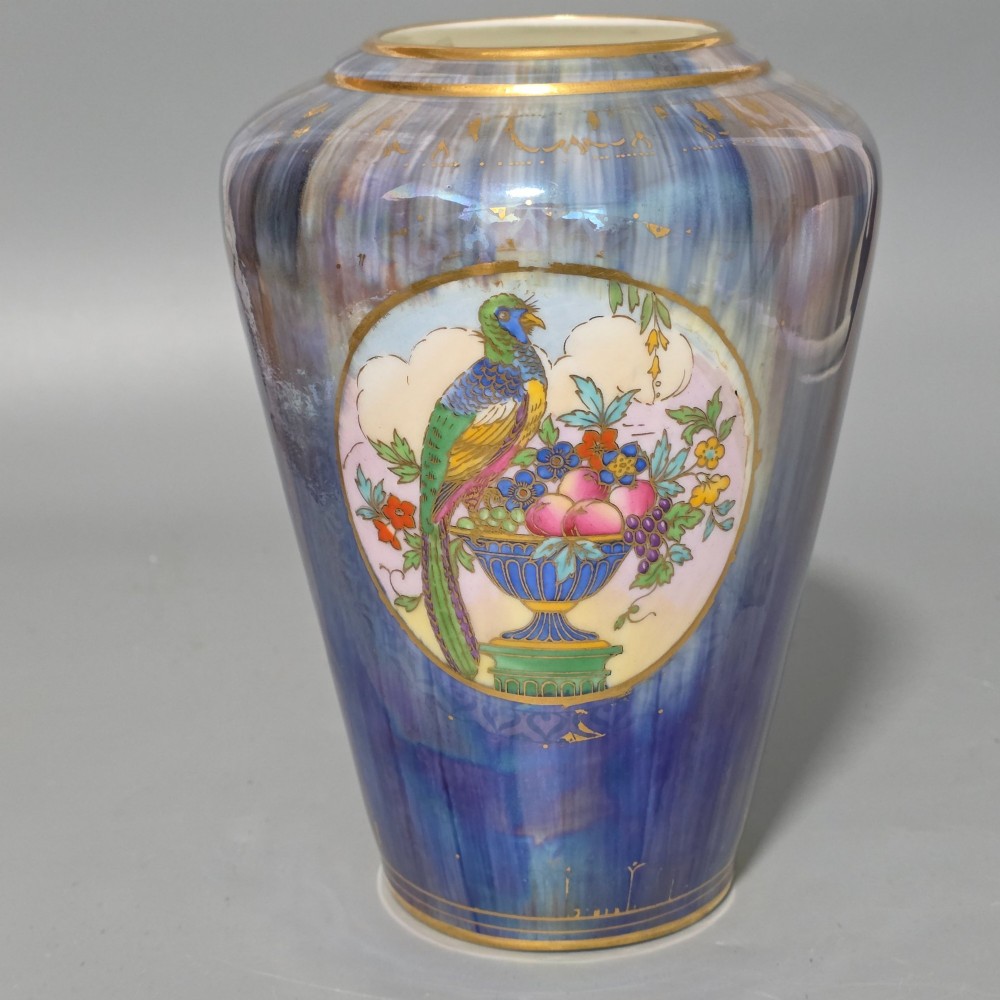 A tapered ceramic vase featuring a streaked iridescent blue lustre glaze and a central circular reserve illustrating a hand-enamelled parrot perched beside a fruit bowl.