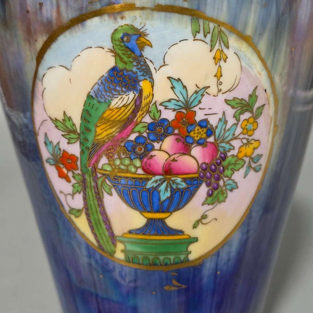 A tapered ceramic vase featuring a streaked iridescent blue lustre glaze and a central circular reserve illustrating a hand-enamelled parrot perched beside a fruit bowl. View IV