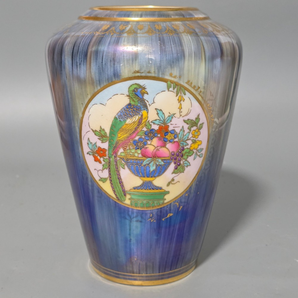 A tapered ceramic vase featuring a streaked iridescent blue lustre glaze and a central circular reserve illustrating a hand-enamelled parrot perched beside a fruit bowl. View V