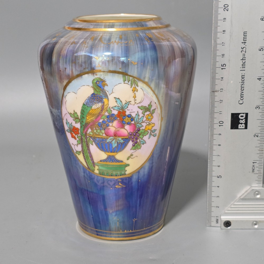 A tapered ceramic vase featuring a streaked iridescent blue lustre glaze and a central circular reserve illustrating a hand-enamelled parrot perched beside a fruit bowl. View VI