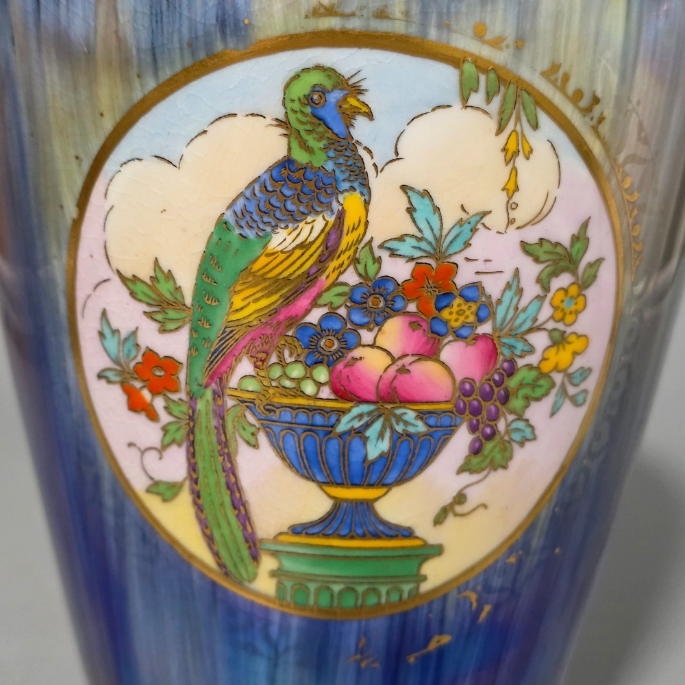 A tapered ceramic vase featuring a streaked iridescent blue lustre glaze and a central circular reserve illustrating a hand-enamelled parrot perched beside a fruit bowl. View IX