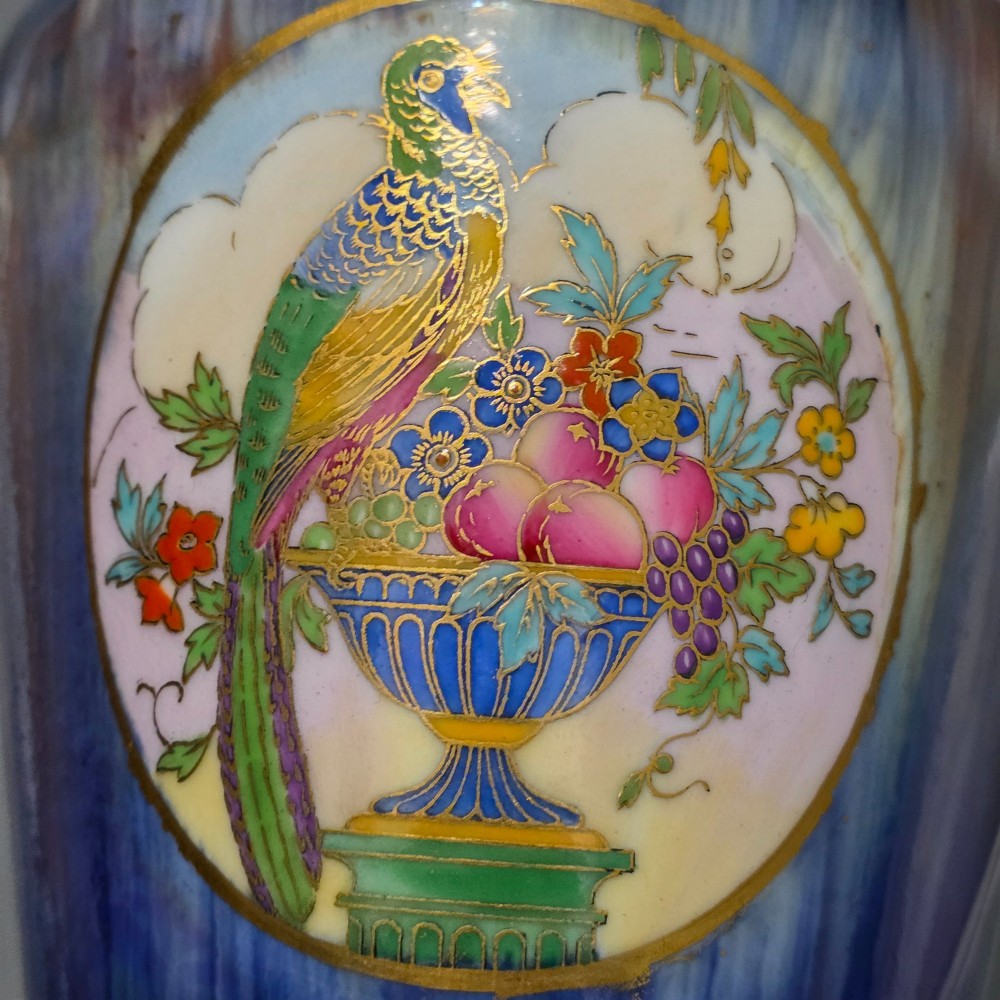 A tapered ceramic vase featuring a streaked iridescent blue lustre glaze and a central circular reserve illustrating a hand-enamelled parrot perched beside a fruit bowl. View XIV