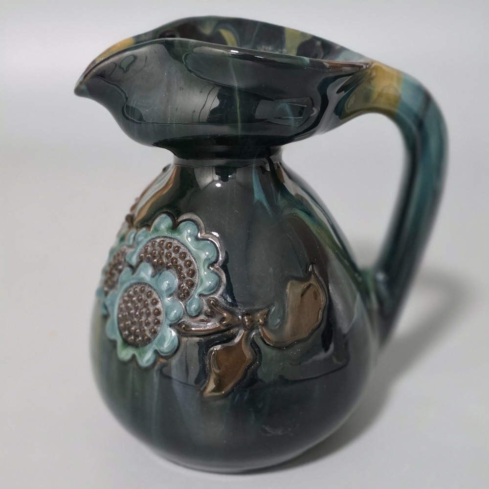 A dark green glazed earthenware jug featuring raised slip floral motifs with blue and brown details, a wide spout, and a single handle.