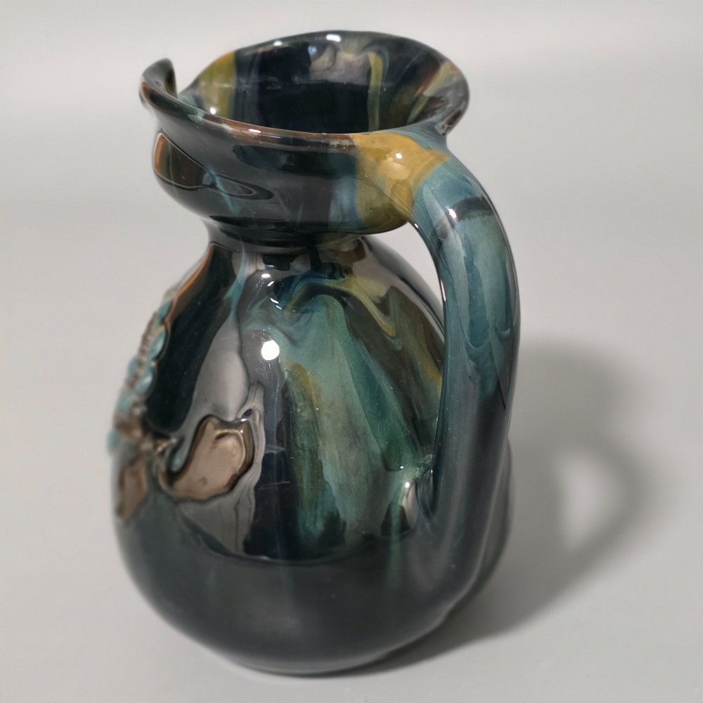 A dark green glazed earthenware jug featuring raised slip floral motifs with blue and brown details, a wide spout, and a single handle. View II