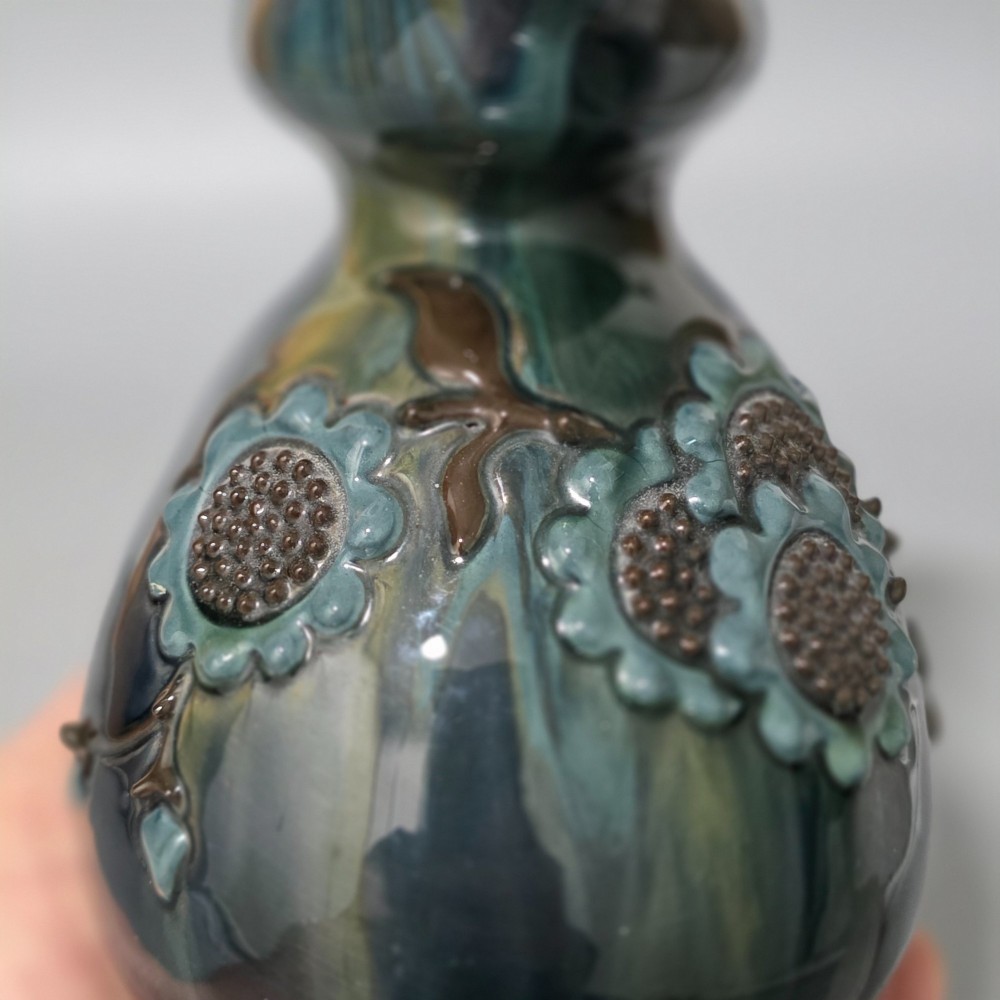 A dark green glazed earthenware jug featuring raised slip floral motifs with blue and brown details, a wide spout, and a single handle. View IV