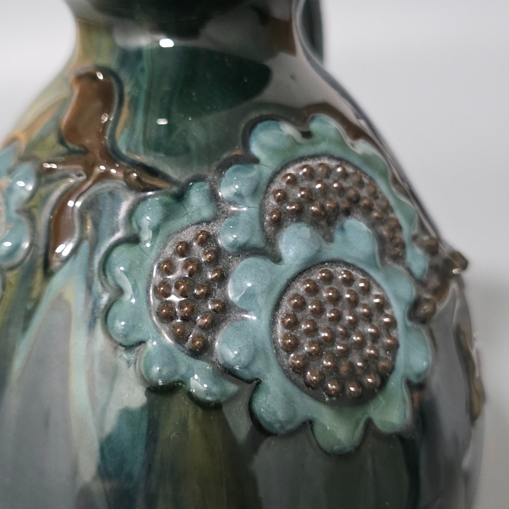 A dark green glazed earthenware jug featuring raised slip floral motifs with blue and brown details, a wide spout, and a single handle. View V