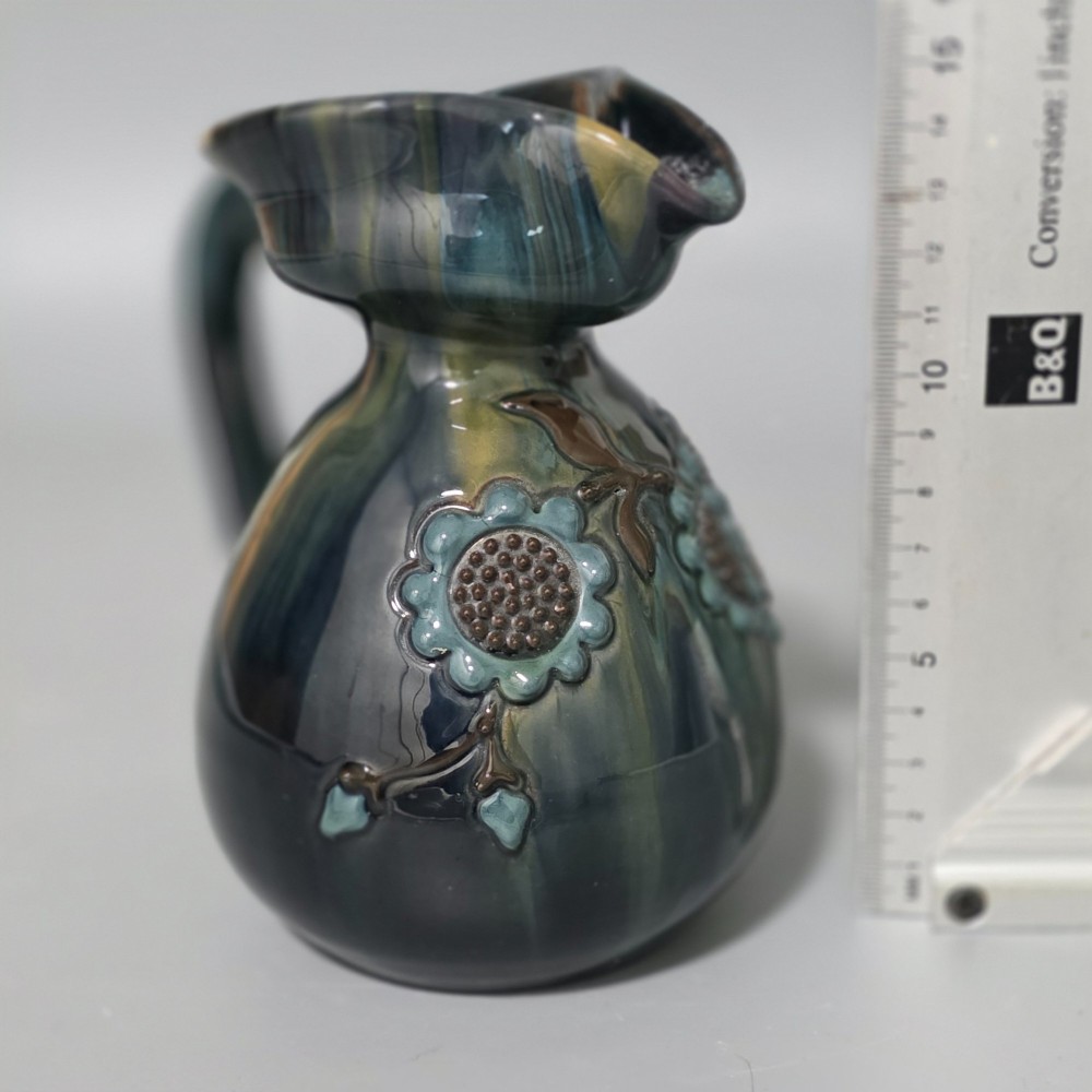 A dark green glazed earthenware jug featuring raised slip floral motifs with blue and brown details, a wide spout, and a single handle. View VI