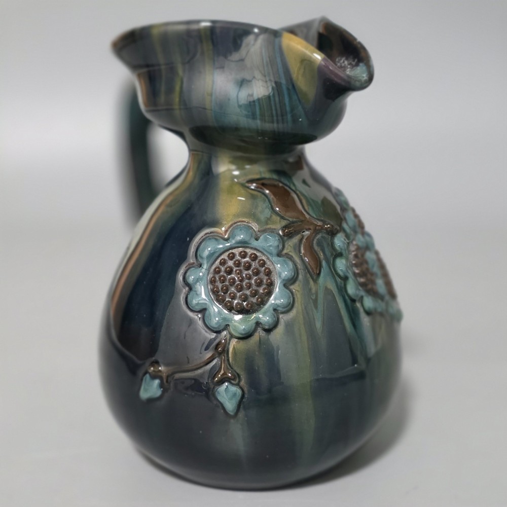 A dark green glazed earthenware jug featuring raised slip floral motifs with blue and brown details, a wide spout, and a single handle. View VII