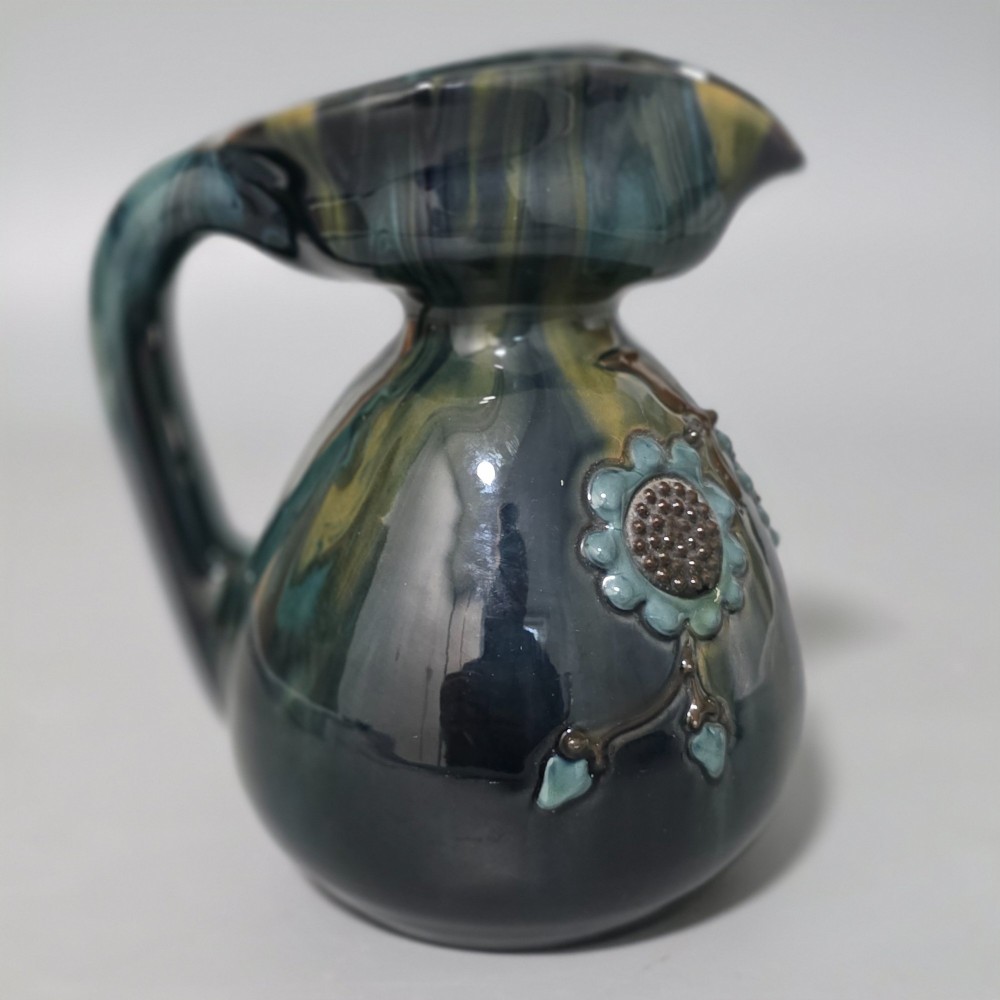A dark green glazed earthenware jug featuring raised slip floral motifs with blue and brown details, a wide spout, and a single handle. View VIII