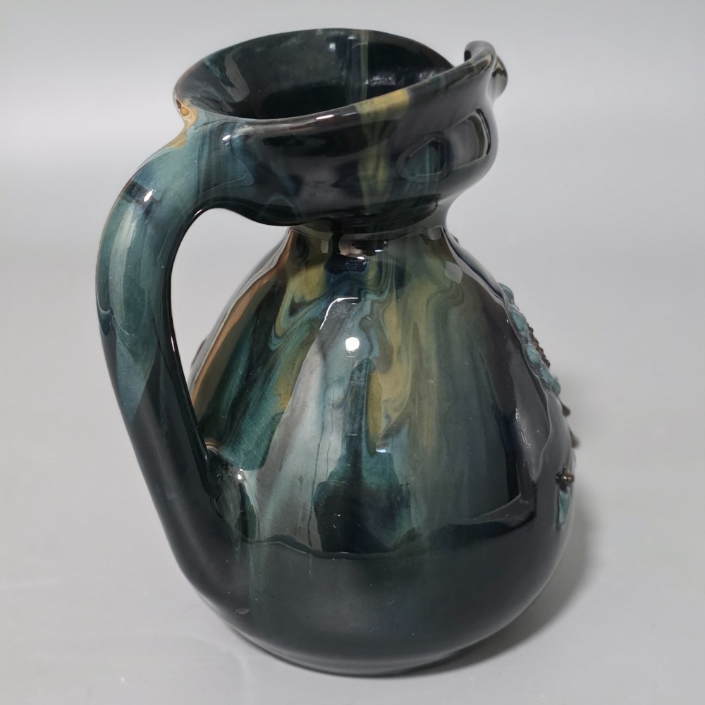 A dark green glazed earthenware jug featuring raised slip floral motifs with blue and brown details, a wide spout, and a single handle. View IX