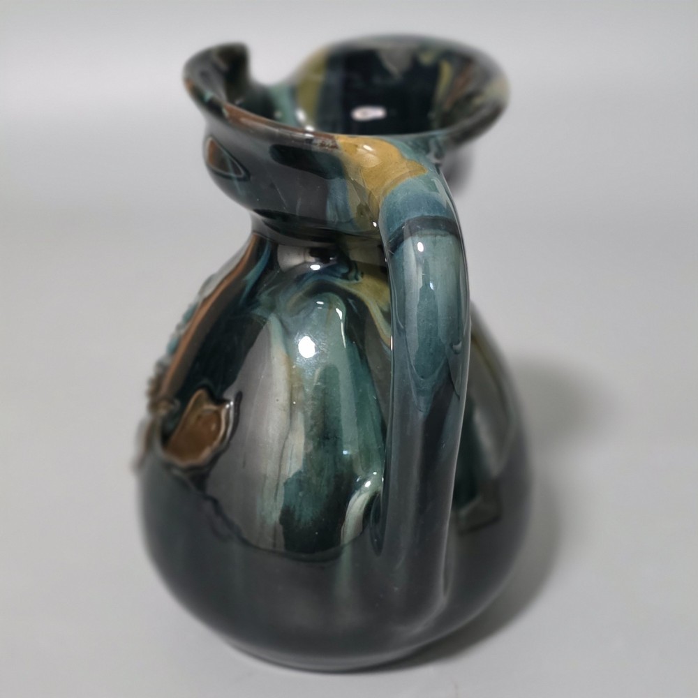 A dark green glazed earthenware jug featuring raised slip floral motifs with blue and brown details, a wide spout, and a single handle. View X