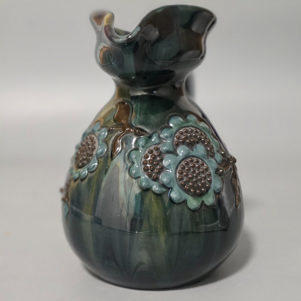 A dark green glazed earthenware jug featuring raised slip floral motifs with blue and brown details, a wide spout, and a single handle. View XI