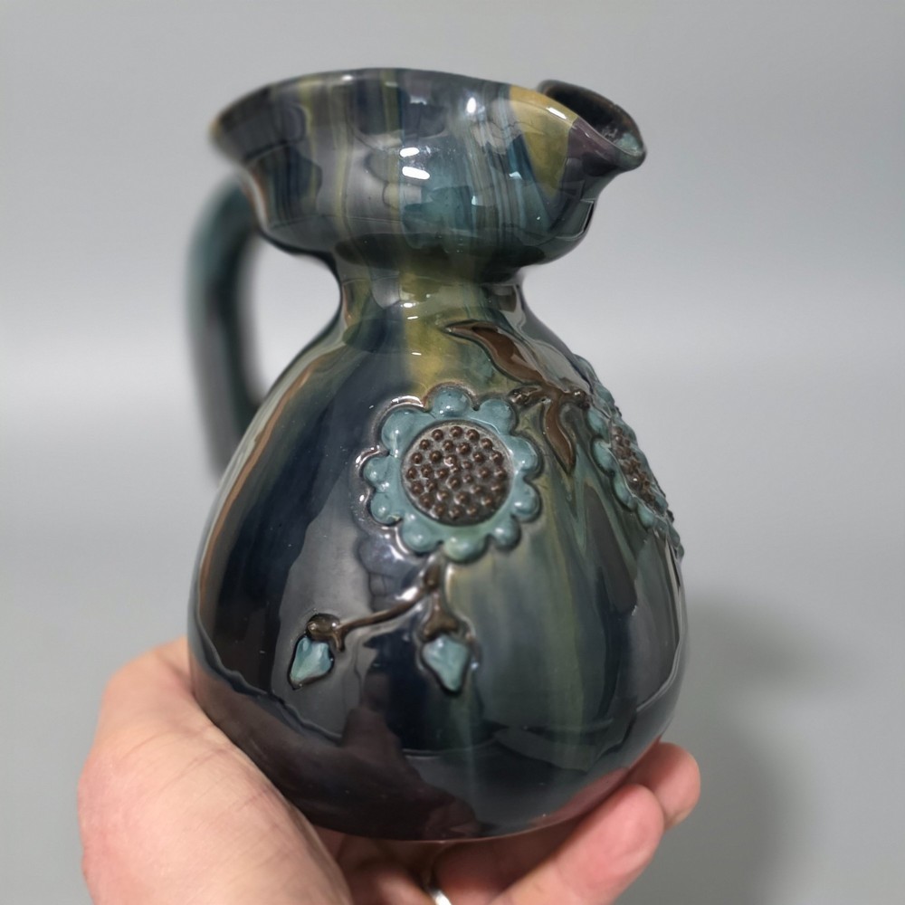 A dark green glazed earthenware jug featuring raised slip floral motifs with blue and brown details, a wide spout, and a single handle. View XII