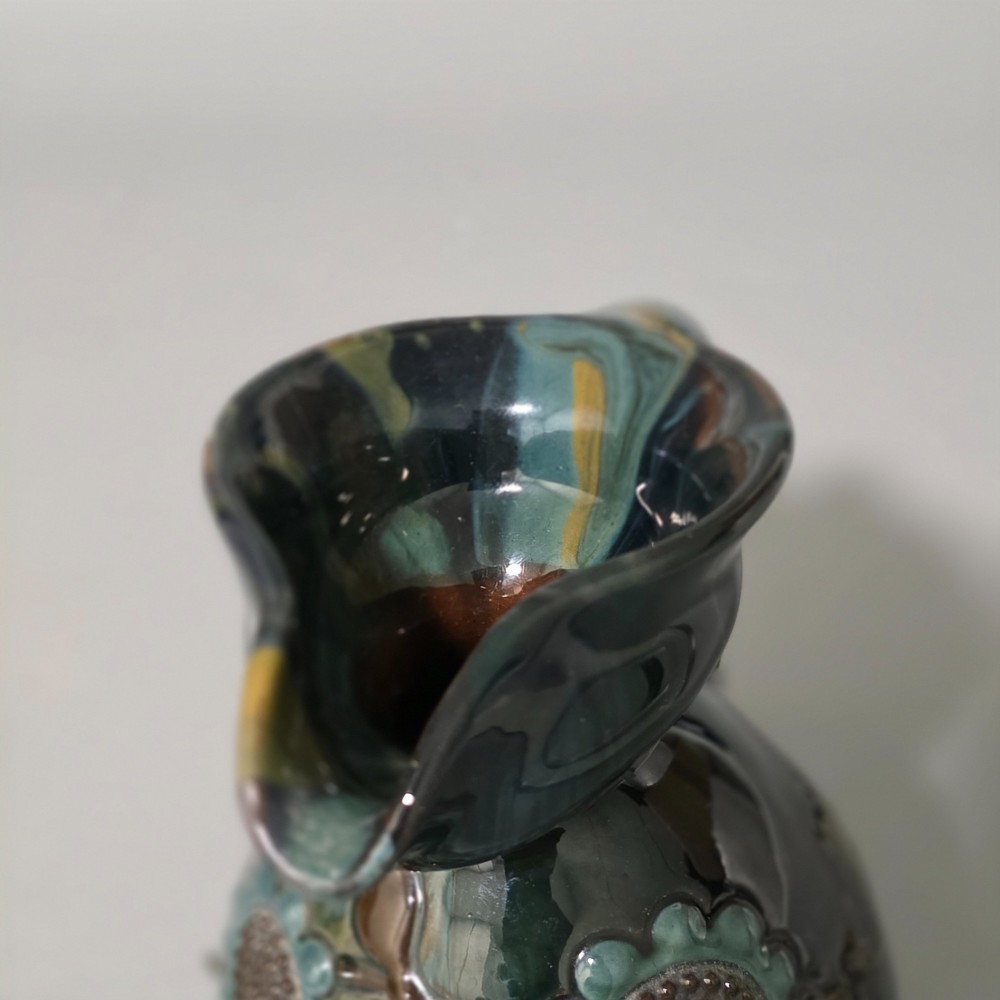 A dark green glazed earthenware jug featuring raised slip floral motifs with blue and brown details, a wide spout, and a single handle. View XIII