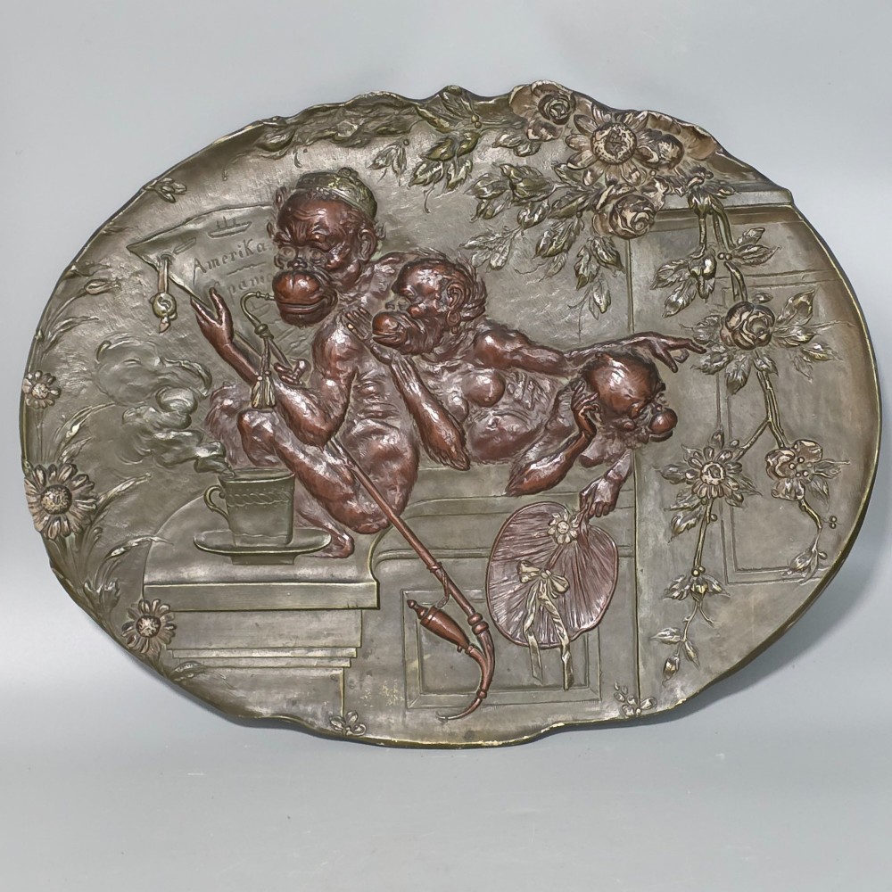 An oval dark brown Austrian terracotta relief plaque depicting a satirical scene of an anthropomorphic monkey family framed by relief floral motifs.