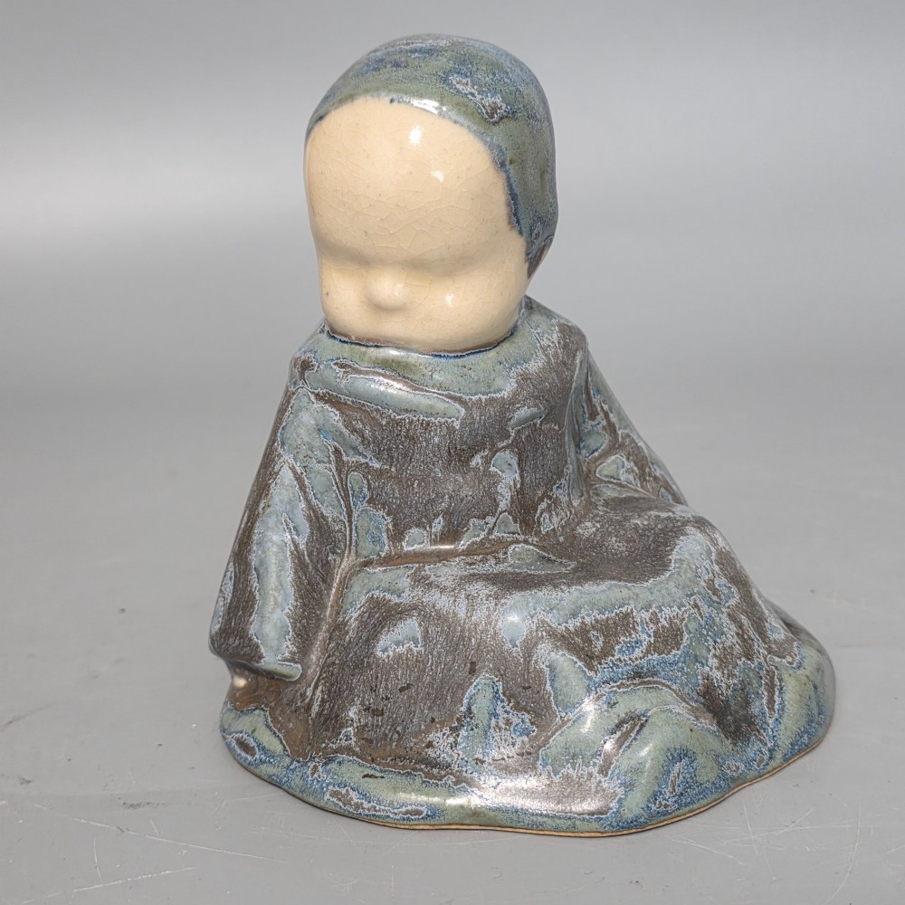 A glazed stoneware figurine depicting a seated child wearing a bonnet, rendered with a mottled blue and grey glaze and a smooth cream face.