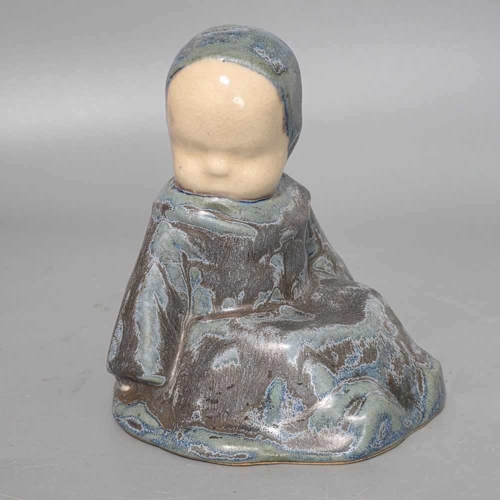 A glazed stoneware figurine depicting a seated child wearing a bonnet, rendered with a mottled blue and grey glaze and a smooth cream face. View II