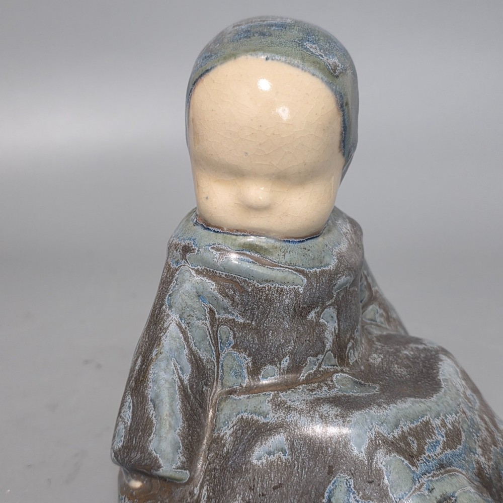 A glazed stoneware figurine depicting a seated child wearing a bonnet, rendered with a mottled blue and grey glaze and a smooth cream face. View III