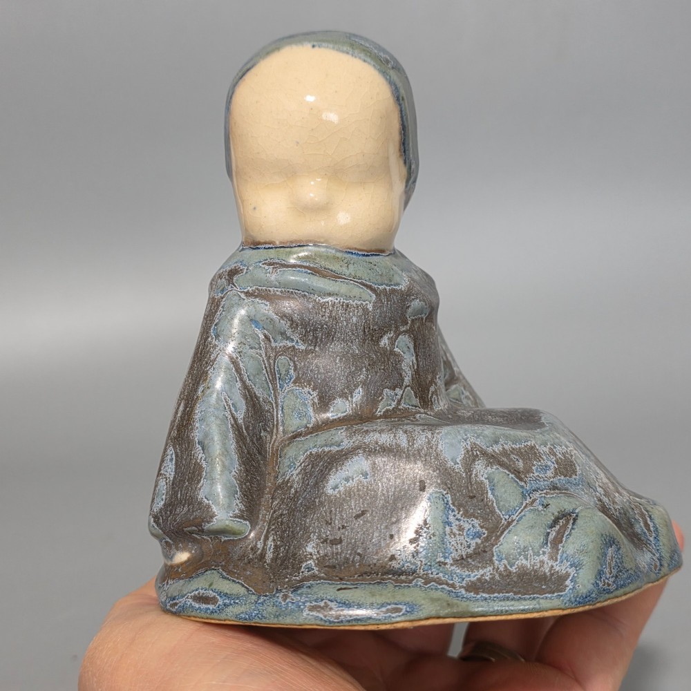A glazed stoneware figurine depicting a seated child wearing a bonnet, rendered with a mottled blue and grey glaze and a smooth cream face. View IV