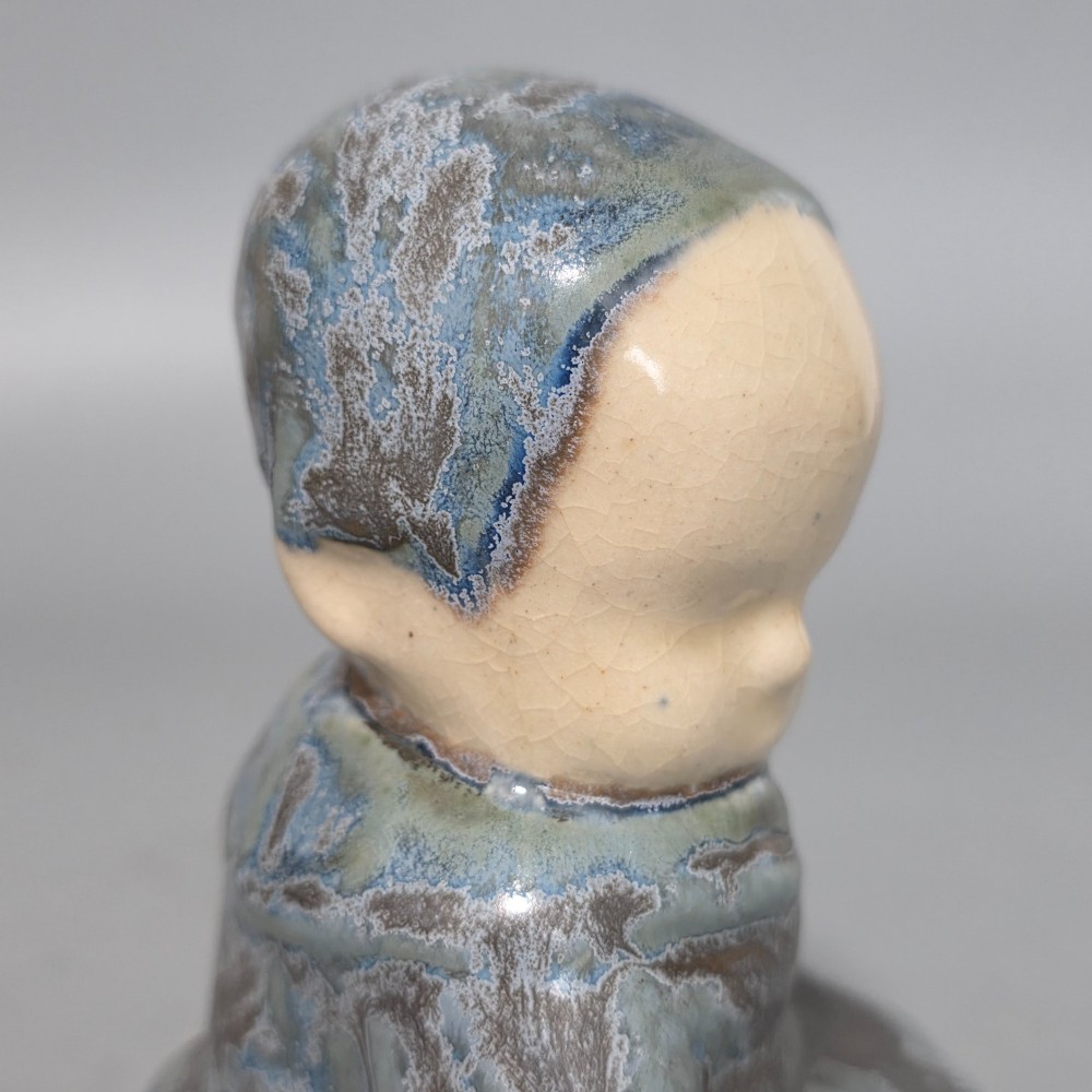 A glazed stoneware figurine depicting a seated child wearing a bonnet, rendered with a mottled blue and grey glaze and a smooth cream face. View V