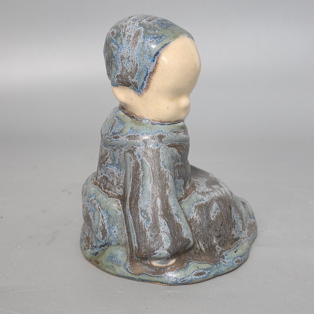 A glazed stoneware figurine depicting a seated child wearing a bonnet, rendered with a mottled blue and grey glaze and a smooth cream face. View IX