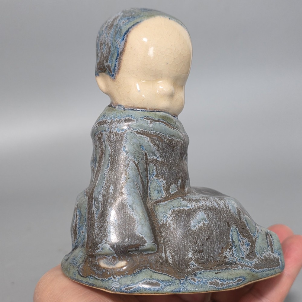 A glazed stoneware figurine depicting a seated child wearing a bonnet, rendered with a mottled blue and grey glaze and a smooth cream face. View X