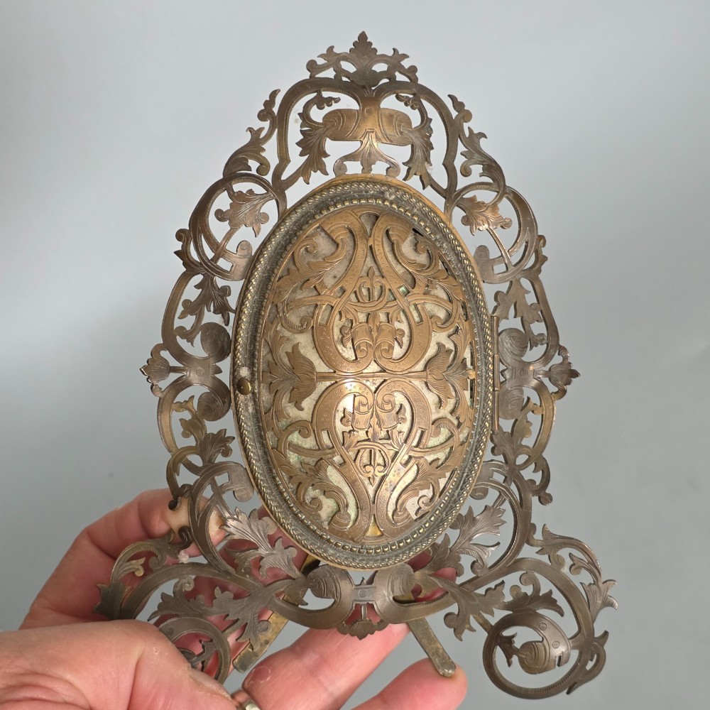 An ornate late Victorian easel photo frame crafted from pierced brass scrollwork with a central hinged oval door. View VII