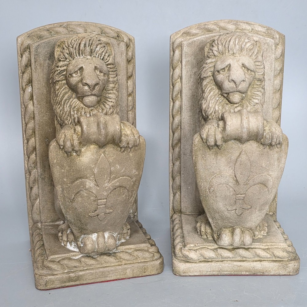 A pair of grey cast stone bookends sculpted as seated lions holding shields featuring a fleur-de-lis motif.