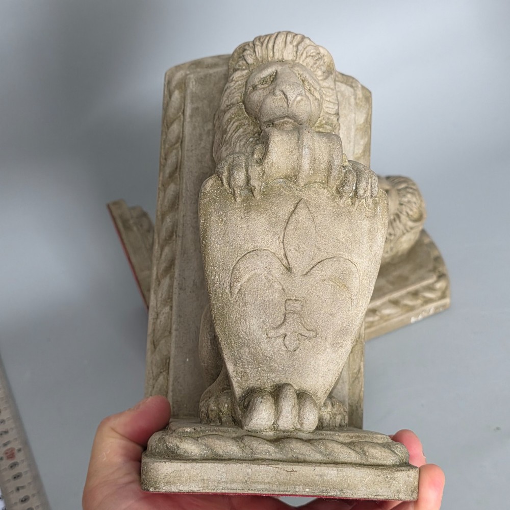 A pair of grey cast stone bookends sculpted as seated lions holding shields featuring a fleur-de-lis motif. View VIII