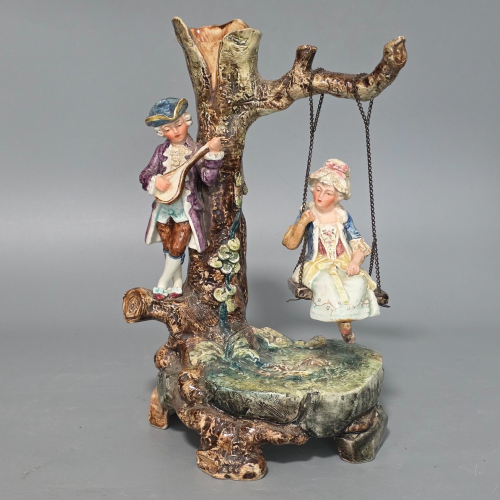 A Victorian majolica spill holder shaped as a hollow tree trunk with a figure of a man playing a lute next to a woman suspended on a swing.