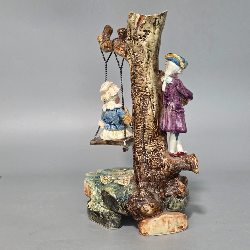 A Victorian majolica spill holder shaped as a hollow tree trunk with a figure of a man playing a lute next to a woman suspended on a swing. View V