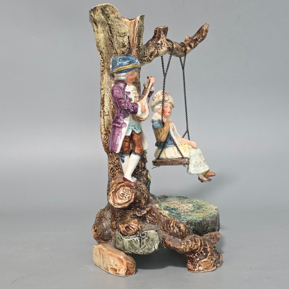 A Victorian majolica spill holder shaped as a hollow tree trunk with a figure of a man playing a lute next to a woman suspended on a swing. View XI