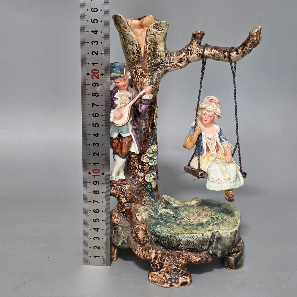 A Victorian majolica spill holder shaped as a hollow tree trunk with a figure of a man playing a lute next to a woman suspended on a swing. View XII