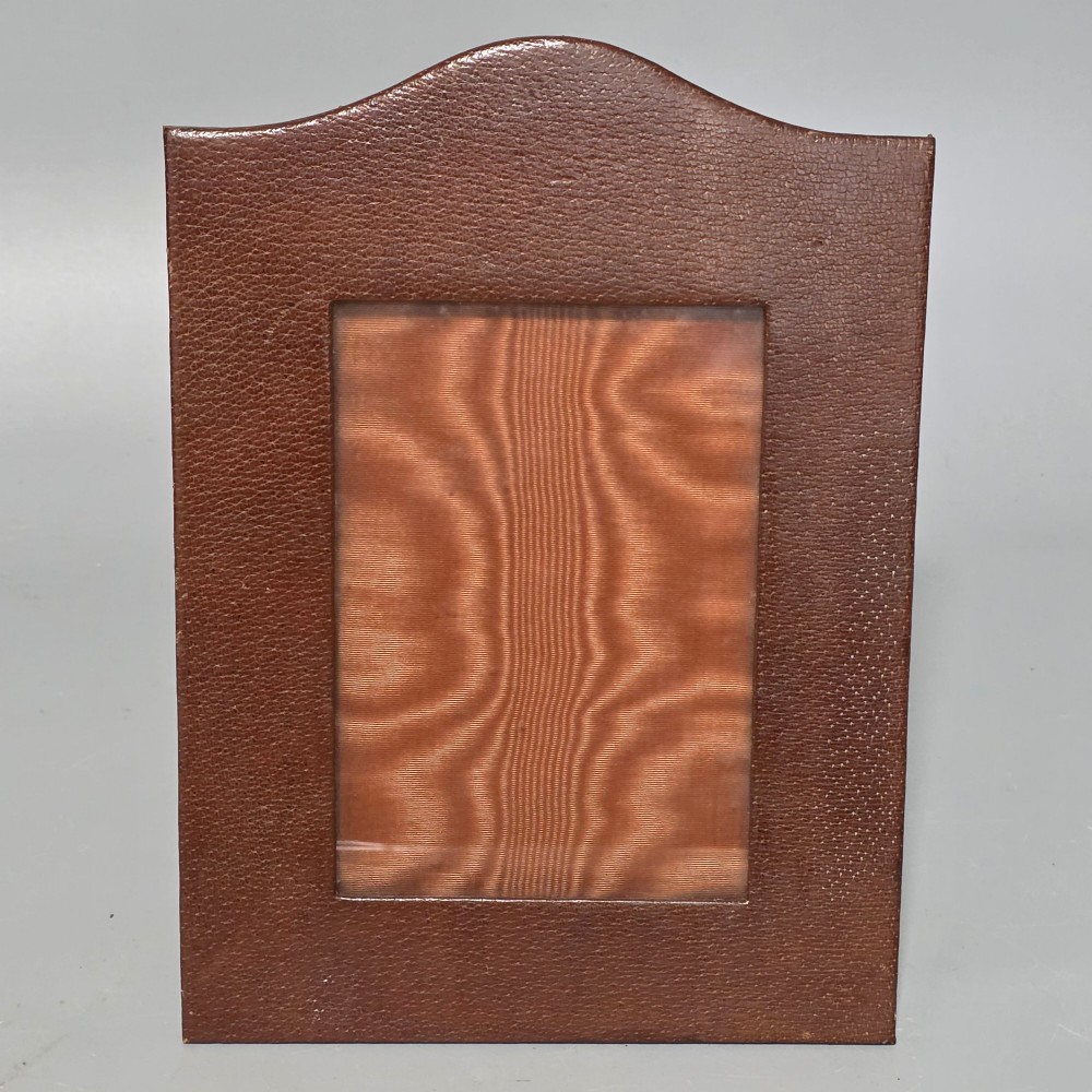 A brown textured leather photo frame featuring an arched top and a central rectangular aperture backed with moire silk.