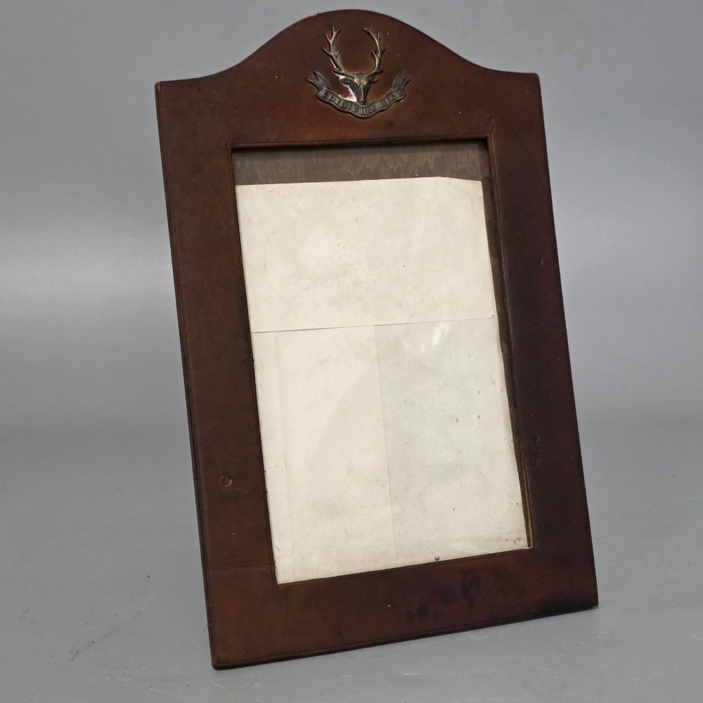 A dark brown leather desktop picture frame featuring an arched top decorated with a metal stag head emblem and motto scroll.