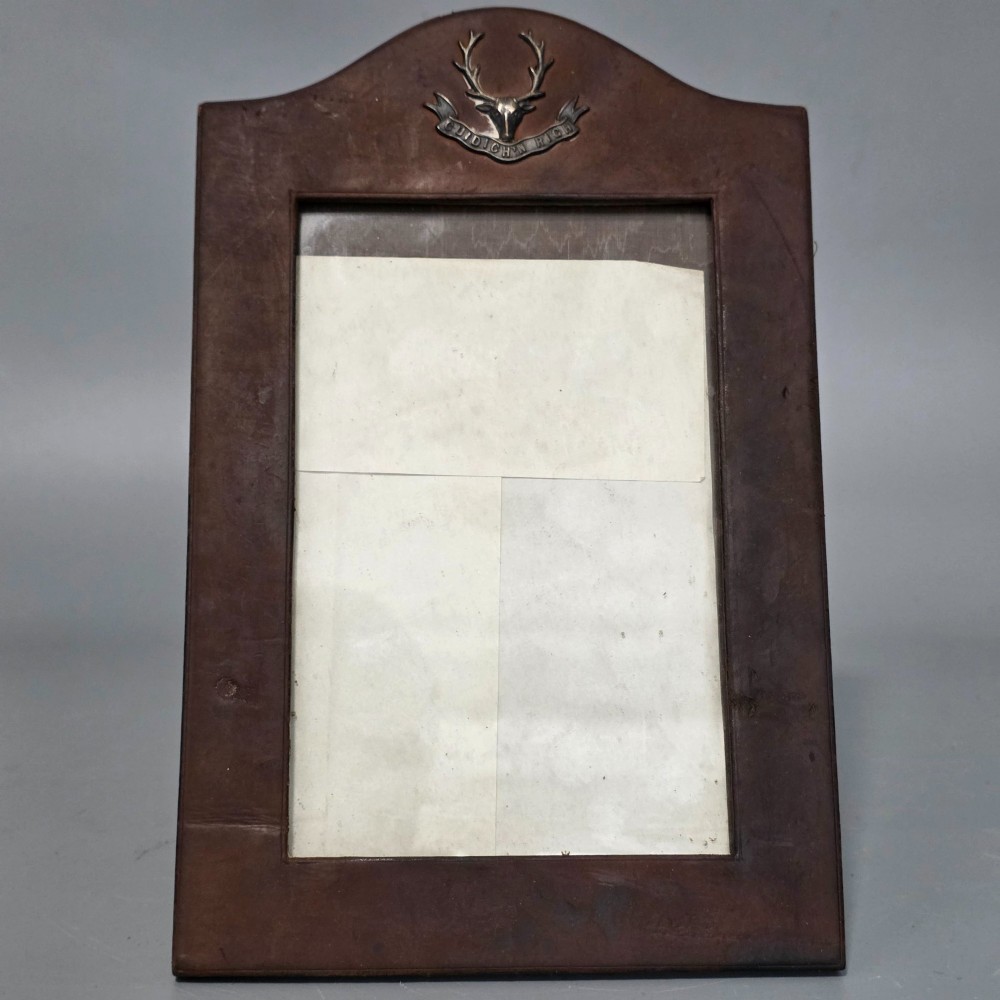 A dark brown leather desktop picture frame featuring an arched top decorated with a metal stag head emblem and motto scroll. View IV