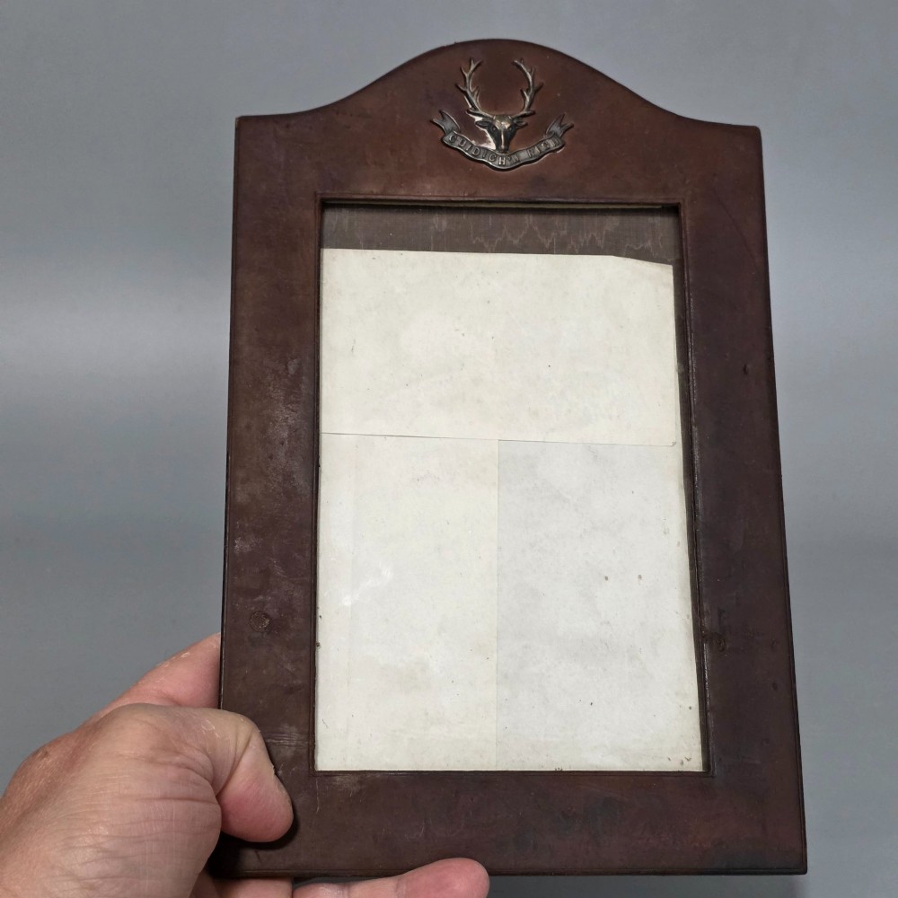 A dark brown leather desktop picture frame featuring an arched top decorated with a metal stag head emblem and motto scroll. View VI