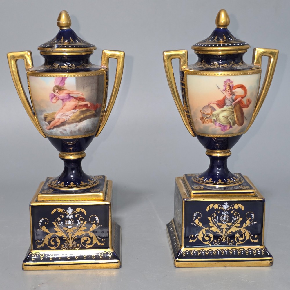A pair of cobalt blue and gold Vienna porcelain Empire amphora vases featuring hand-painted classical figures and square plinth bases.