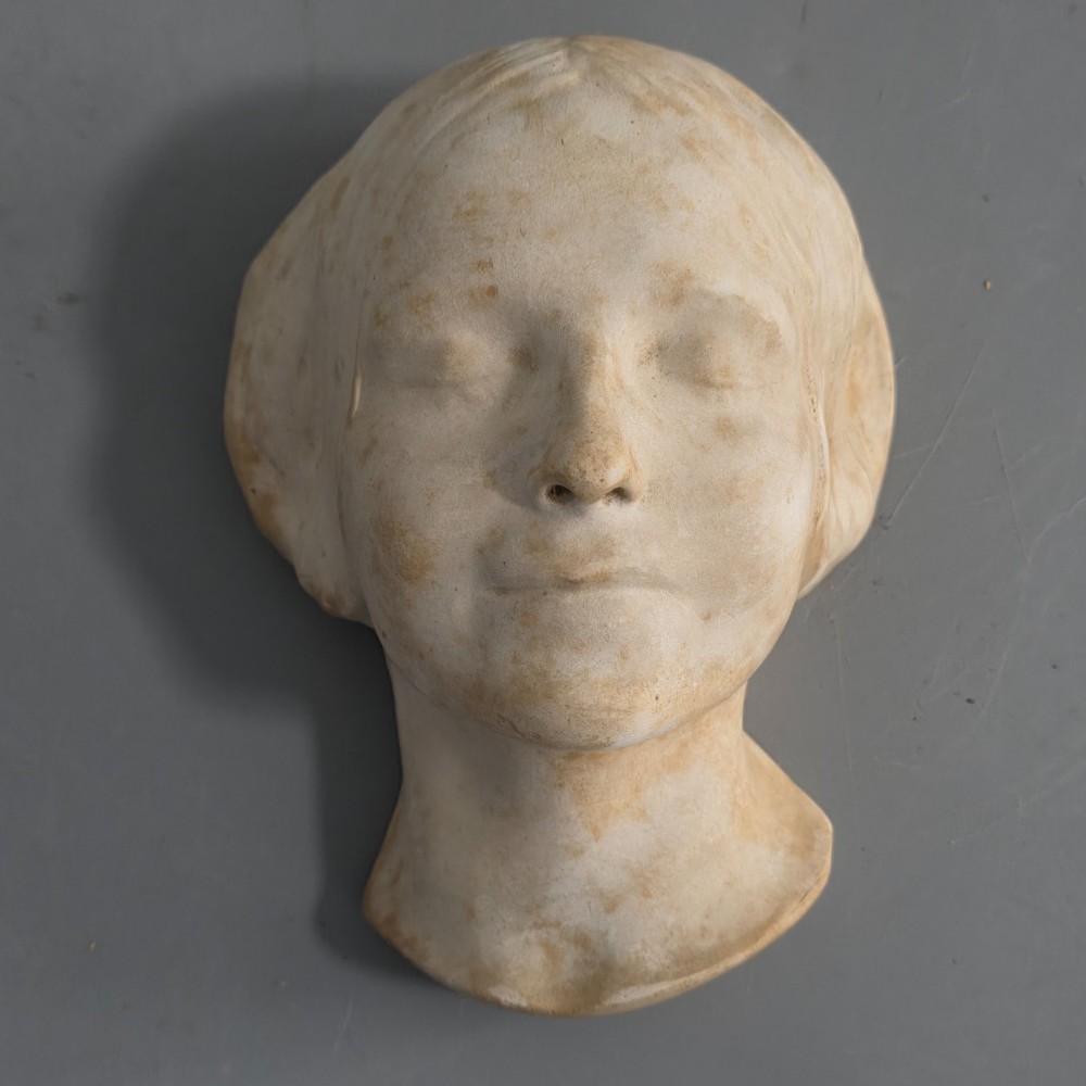 A carved stone death mask depicting a young woman with closed eyes, centre-parted hair, and a subtle smile against a neutral backdrop.
