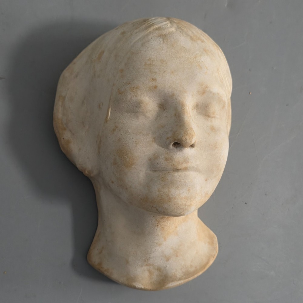 A carved stone death mask depicting a young woman with closed eyes, centre-parted hair, and a subtle smile against a neutral backdrop. View II