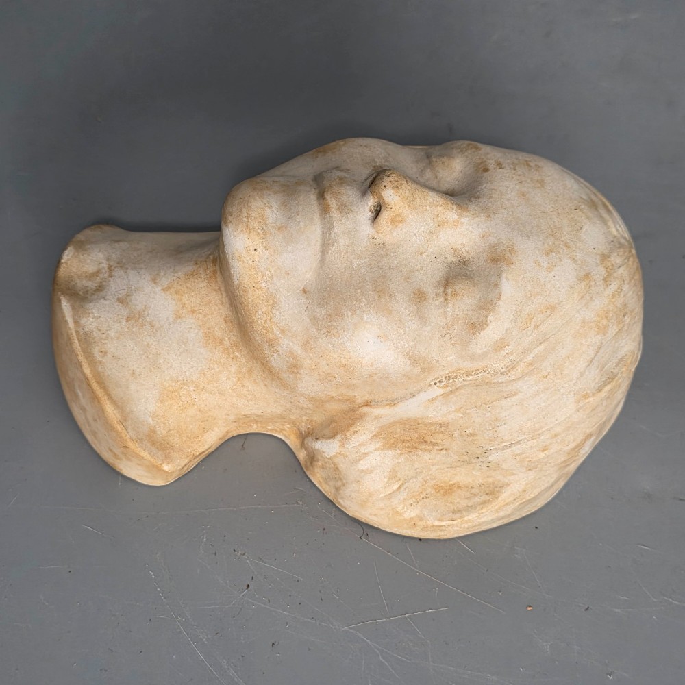 A carved stone death mask depicting a young woman with closed eyes, centre-parted hair, and a subtle smile against a neutral backdrop. View III