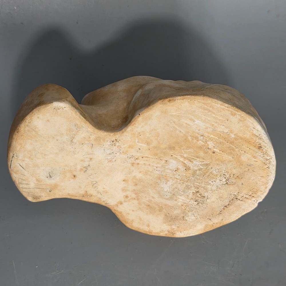 A carved stone death mask depicting a young woman with closed eyes, centre-parted hair, and a subtle smile against a neutral backdrop. View IV