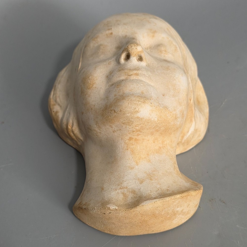 A carved stone death mask depicting a young woman with closed eyes, centre-parted hair, and a subtle smile against a neutral backdrop. View V