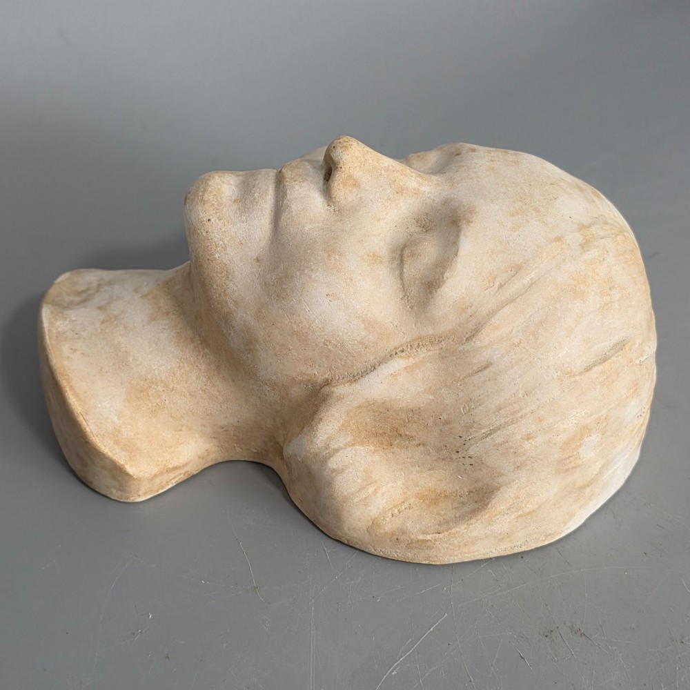 A carved stone death mask depicting a young woman with closed eyes, centre-parted hair, and a subtle smile against a neutral backdrop. View VI