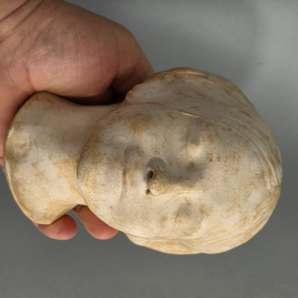 A carved stone death mask depicting a young woman with closed eyes, centre-parted hair, and a subtle smile against a neutral backdrop. View VII