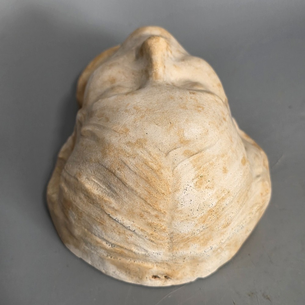 A carved stone death mask depicting a young woman with closed eyes, centre-parted hair, and a subtle smile against a neutral backdrop. View IX