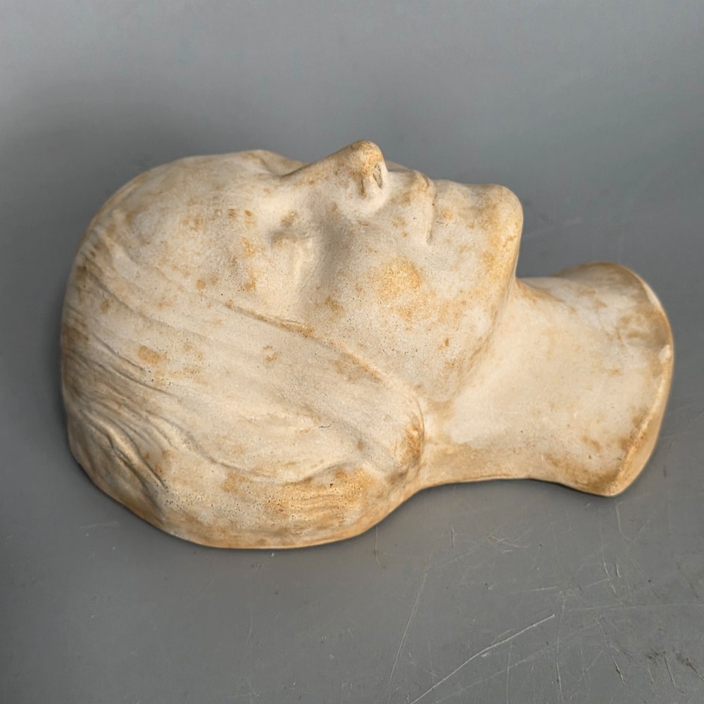 A carved stone death mask depicting a young woman with closed eyes, centre-parted hair, and a subtle smile against a neutral backdrop. View X