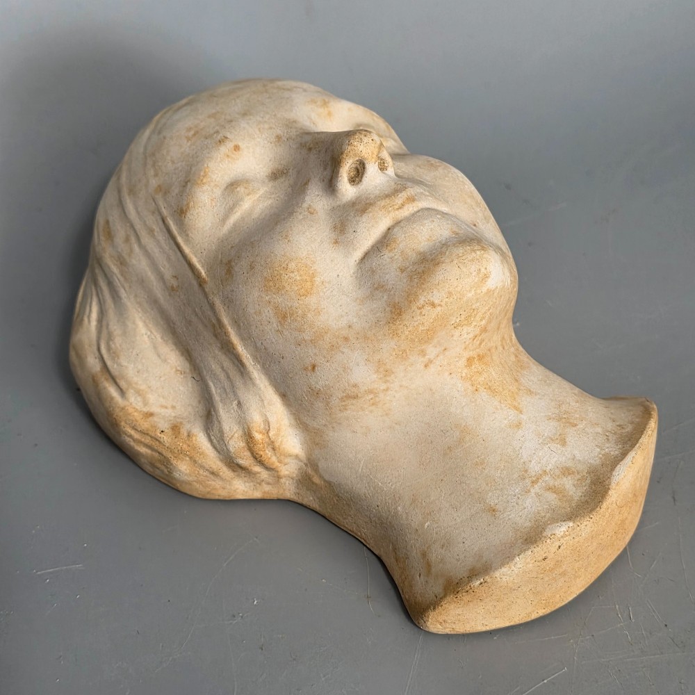 A carved stone death mask depicting a young woman with closed eyes, centre-parted hair, and a subtle smile against a neutral backdrop. View XI