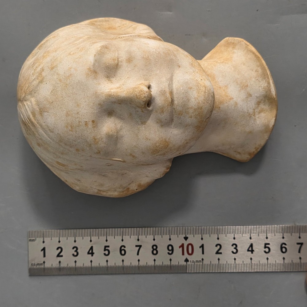 A carved stone death mask depicting a young woman with closed eyes, centre-parted hair, and a subtle smile against a neutral backdrop. View VIII
